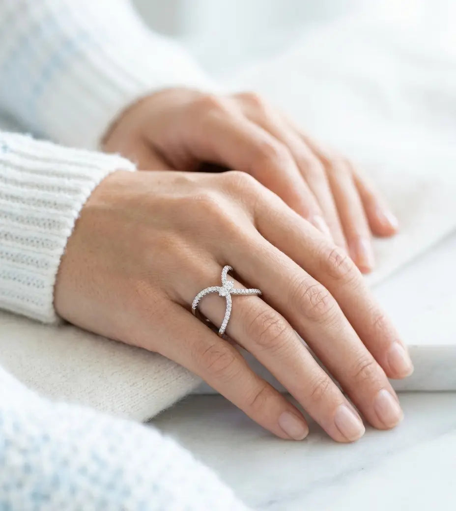 A close-up of a person's hands resting on a white surface, wearing a delicate silver ring with small clear stones in an X-shaped design. The person is wearing a white ribbed knit sweater.